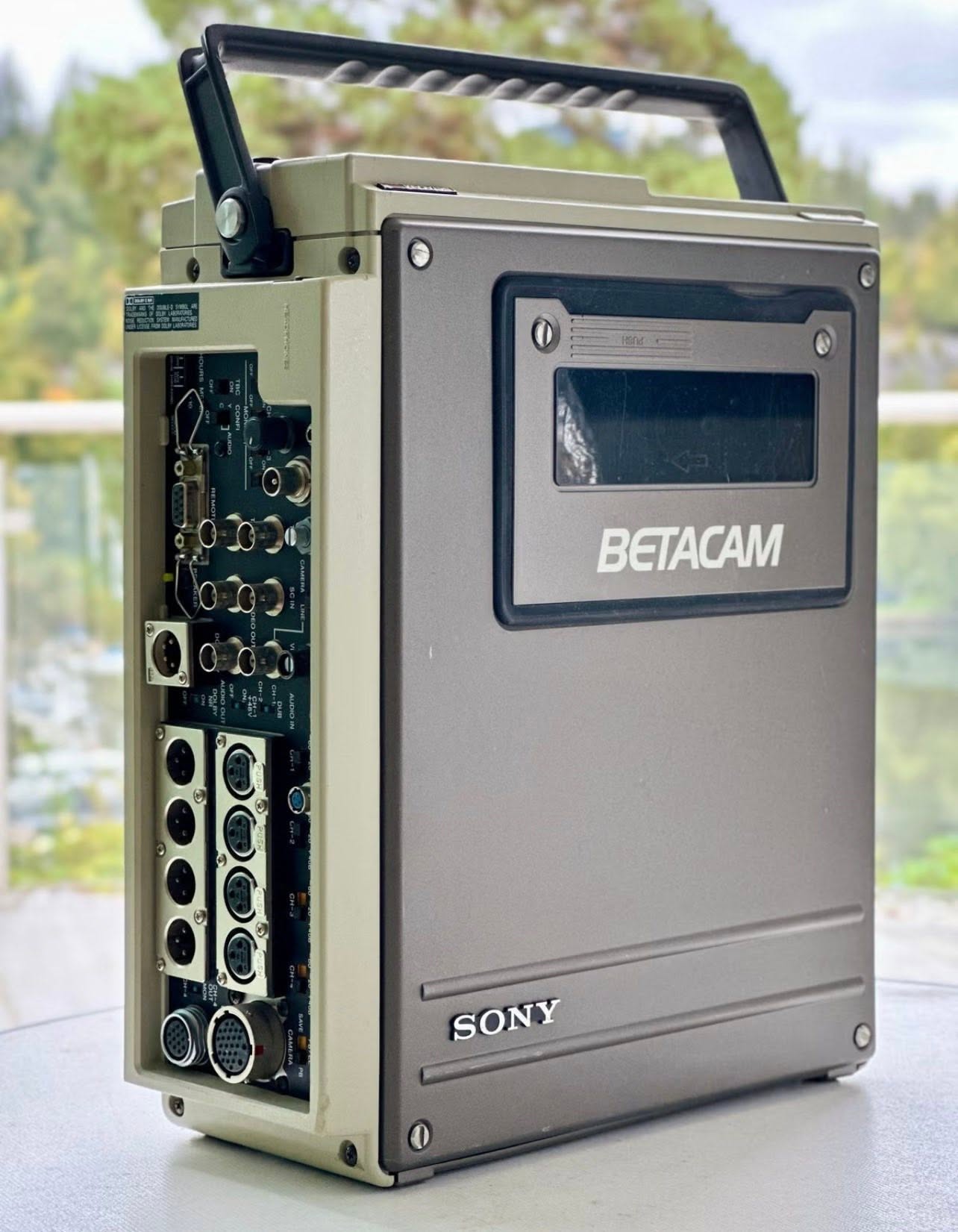 Sony BETACAM videokassettopptaker med åpen tilkoblingspanel og skjerm, stående utendørs mot grønn bakgrunn.